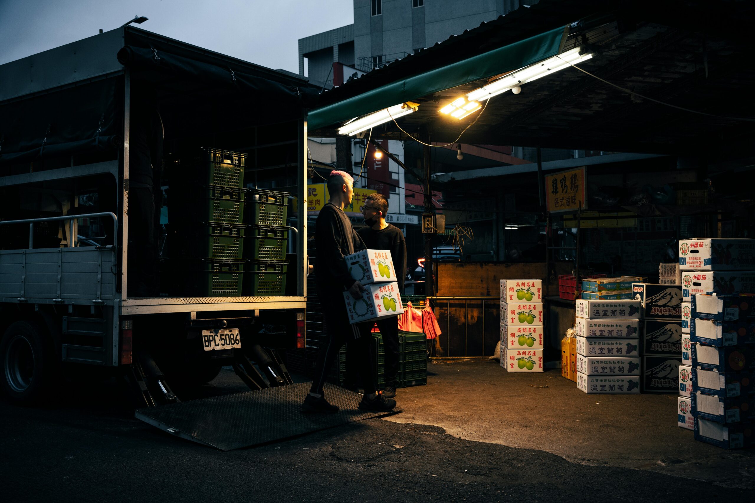 Workers unloading boxes from a delivery truck at an urban market during dusk.